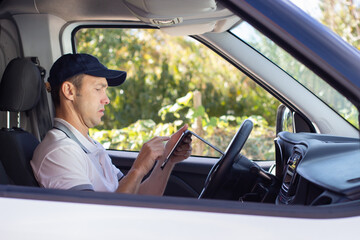 Side view of deliveryman checking mobile phone in car. Mid adult man in white T-shirt and cap...