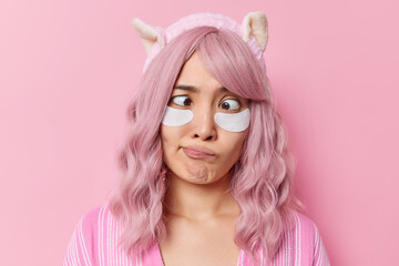 Naklejka premium Horizontal shot of rosy haired Asian woman crosses eyes applies beauty patches for skin treatment has dyed pink hair wears soft headband poses against rosy background takes care of her complexion