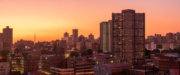 A horizontal panoramic cityscape taken after sunset, against a pink and orange sky, of the central business district of the city of Johannesburg, South Africa