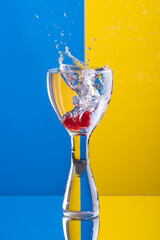 A vertical studio shot of a red cherry being dropped from a height into clear liquid in a cocktail glass against a blue and yellow background, creating a splash of liquid. Johannesburg, South Africa