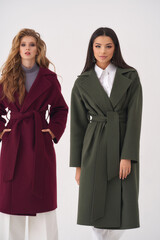 Attractive long haired young women blonde and brunette wearing colorful warm woolen coats with tied belts stand together in white studio