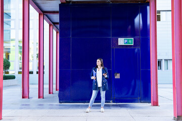 Beautiful Caucasian businesswoman waiting with a folder and a phone outside an office building. She is wearing a casual blue jacket.