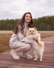 Young woman in oversize knitted woolen sweater holds German Spitz dog standing on wooden footbridge...
