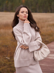 Smiling brunette woman in warm oversize sweater and skirt holds white handbag standing on wooden footbridge at countryside in autumn