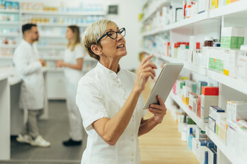 Beautiful senior female pharmacist checking medications on a shelf - obrazy, fototapety, plakaty