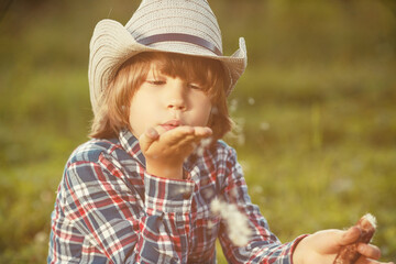 Child in backlight of sunset blows on fluff. Kid is playing dandelion. Boy in cowboy hat and plaid shirt.