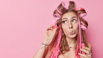 Naklejka premium Beautiful young woman applies mascara on eyelashes makes daily makeup wears hair curlers purses lips concentrated away isolated over pink background blank copy space on right gets ready fo party