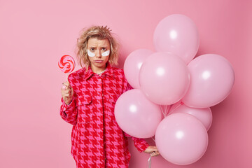 Obraz na płótnie Canvas Frustrated upset woman has bad mood after party holds delcious round candy on stick and bunch of inflated airballoons wears beauty patches dressed in fashionable clothes isolated over pink wall