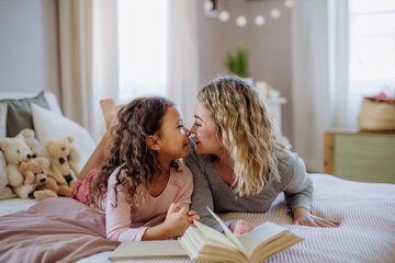 Happy mother with her little daughter lying on bed and tlooking at each other when reading book.