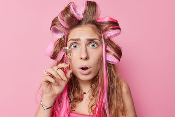 Headshot of embarrassed young blue eyed young European woman uses eyelashes curler stares bugged eyes wears hair rollers stands against pink background feels worried being in hurry for date.