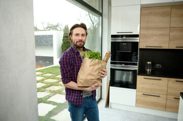 Fototapeta premium Handsome Caucasian man, charming guy, stands in a home kitchen with a cardboard bag full of healthy food and whole grain bread, looks at camera against the background of windows overlooking the garden