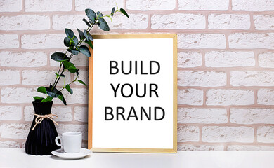 On a white table against a light brick wall, a branch of eucalyptus in a dark vase, a white cup and a light wooden frame with the text BUILD YOUR BRAND. Home office interior.