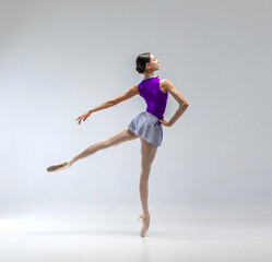 Young and beautiful ballerina in purple ballet leotard and grey skirt. She wears ballet pointe shoes and dancing in white studio. - obrazy, fototapety, plakaty