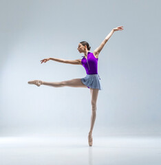 Young and beautiful ballerina in purple ballet leotard and grey skirt. She wears ballet pointe shoes and dancing in white studio.