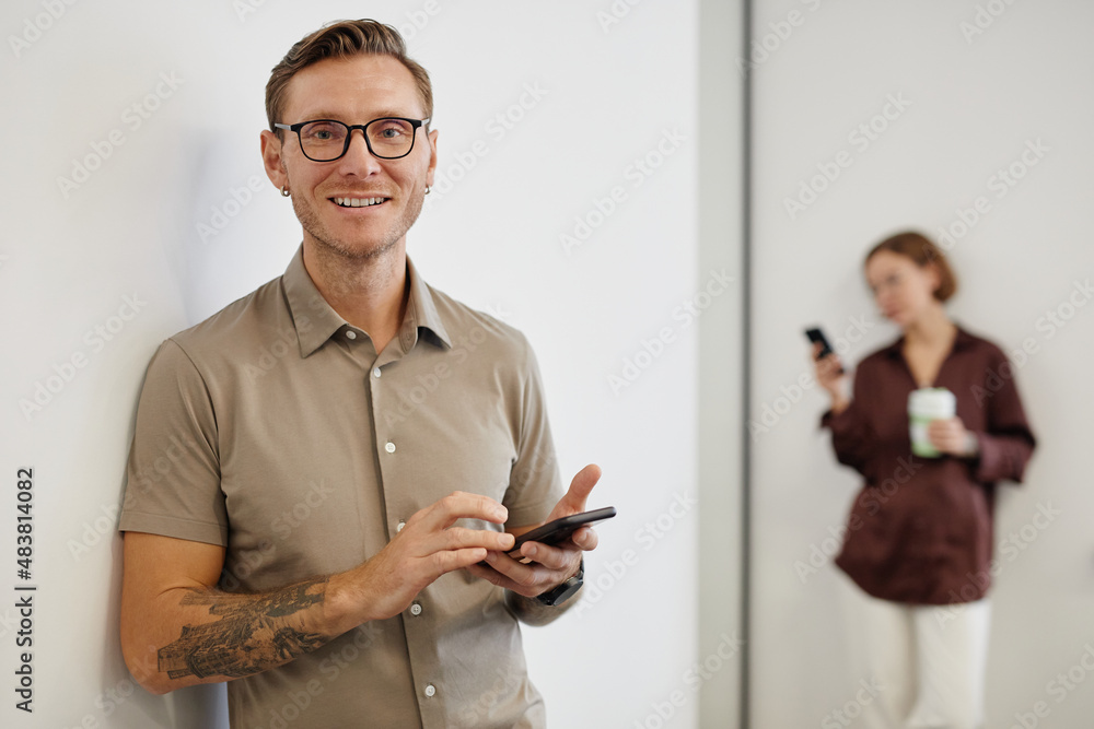 Wall mural minimal waist up portrait of smiling tattooed man holding smartphone and looking at camera in office