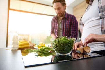 Focus on a glass bowl with green lettuce leaves and greens on a kitchen island against the background of a happy pregnant couple slicing fresh vegetables with falling sun rays through large windows