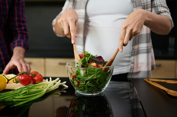 Focus on pregnant woman's hands holding wooden spoons and mixing ingredients in a glass bowl, preparing delicious healthy salad for dinner in the kitchen island next to her husband chopping vegetables