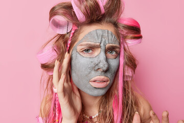 Portrait of displeased woman touches face applies beauty clay mask frowns face wears hair rollers feels unhappy takess care of complexion isolated over pink background. Anti aging procedures