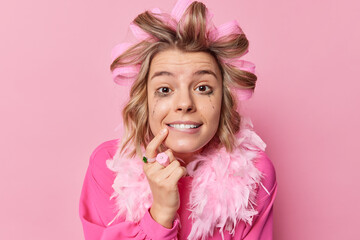 Headshot of cheerful woman with spoiled makeup applies hair curlers prepares for date wears festive outfit bits lips keeps index finger on chin isolated over pink background. Hairstyling concept