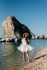 A beautiful young woman in a hat, glasses and a light dress is walking along the ocean shore against the background of huge rocks on a sunny day. Tourism and tourist trips.