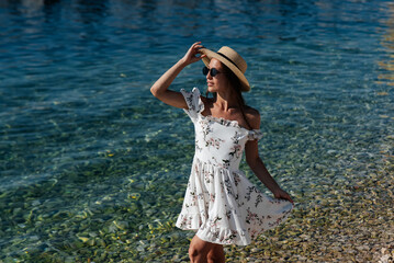 A beautiful young woman in a hat, glasses and a light dress is walking along the ocean shore against the background of huge rocks on a sunny day. Tourism and tourist trips.