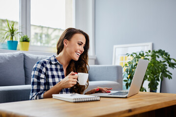 Happy young woman drinking coffee using laptop at home