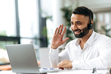 Sociable attractive smart Indian entrepreneur, business man, sales manager, trader, with headphones sit at table in the office, communicates via video call with colleagues or clients, greeting, smiles