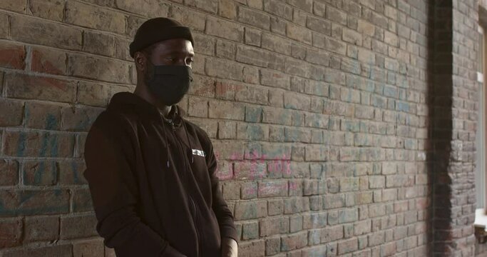young black man in black mask standing at street waiting for victim to commit a crime. stranger guy in hat and black wear. cruel man outdoors at daytime. Concept portrait of dangerous criminal