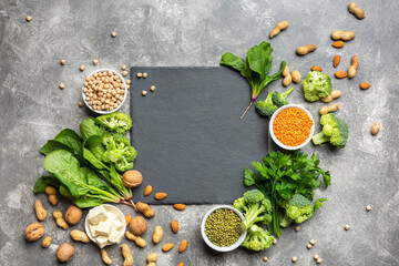 Fototapeta na wymiar A source of protein for vegetarians. Healthy clean food: greens, vegetables, nuts and legumes top view on a concrete background with a black cutting stone in the center.
