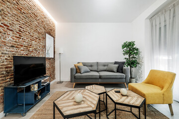 Living room with gray sofa, tv, circular glass dining table, white curtains, yellow personal sofas and blue metal tv table and low polyhedral tables and exposed brick wall