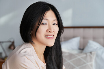 Close up attractive smiling asian woman in casual clothes sitting on the bed at home