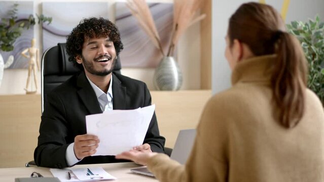 Confident successful Indian male HR manager, entrepreneur, conducts interview girl in a modern office, wearing formal clothes, looking and listening to a job candidate, holding a resume in hands