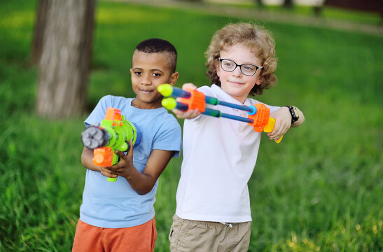 two children with toy guns are smiling in a park against a background of greenery.