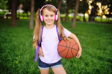 cute little girl in pink headphones and with a basketball is smiling against the background of greenery and a park.