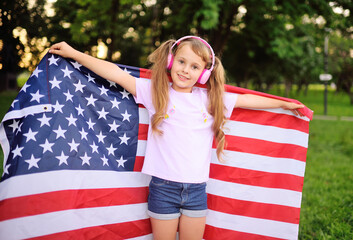 a little girl in denim shorts and pink headphones smiles and holds a large flag of the United...