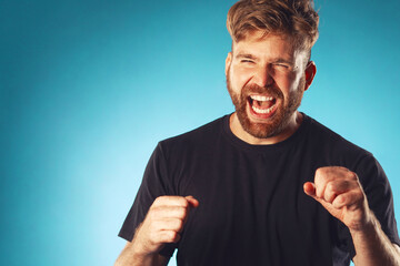 Victory concept. Emotive portrait of happy screaming charismatic active 30-year-old man posing over blue background with hands up. Copy space. Close up studio shot