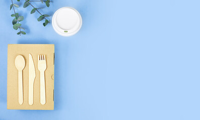 top view of take away box, wooden cutlery and take away cup with green leaves on blue background with copy space