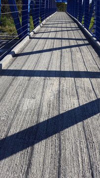 The long concrete strip of a pedestrian crossing or walkover. There are blue decorative pieces of iron and industrial steal on either side which cast shadows on the floor. Some greenery can be seen in