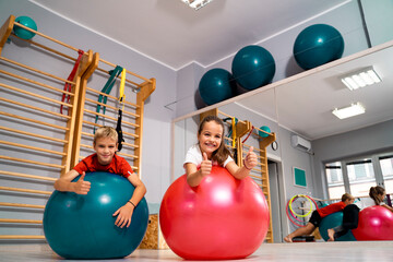Toddlers boy and girl laying on swiss balls in the gym and showing thumbs up, active kids having...