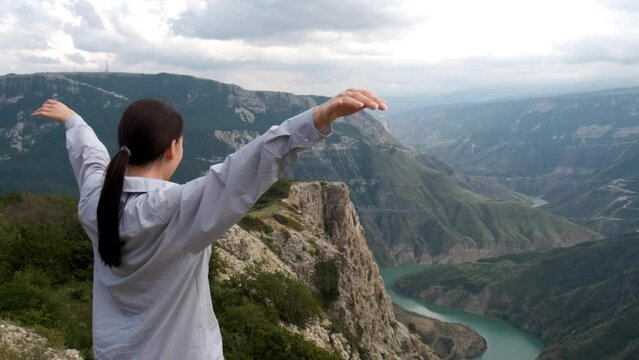 A caucasian brunette girl stands on the edge of a mountain cliff, looks forward, raises her hands up and smiles. Sulak Canyon, Dagestan. Tourism, travel