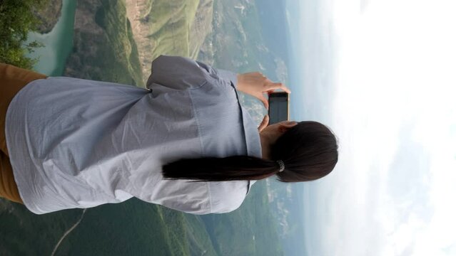 A Caucasian brunette blogger woman stands on the edge of a mountain cliff and shoots a video on her smartphone. Vertical frame. Sulak Canyon, Dagestan. Tourism, travel