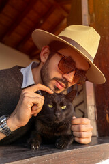 Latin hipster leaning out the window of a traditional colonial house petting his pet a black cat