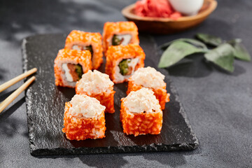Maki sushi on dark stone table. California maki with tobiko. Sushi roll with salmon, cucumber inside, maguro, crab outside. Style concept japanese menu with black background, leaves and hard shadow.