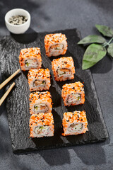 Maki sushi on dark slate. California maki with masago and sesame. Sushi roll with crab, cucumber inside, tobiko outside. Style concept japanese menu with black background, leaves and hard shadow.