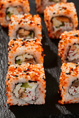 Maki sushi on dark slate. California maki with masago and sesame. Sushi roll with crab, cucumber inside, tobiko outside. Style concept japanese menu with black background, leaves and hard shadow.