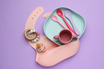 Set of plastic dishware, silicone bib and baby accessories on violet background, flat lay