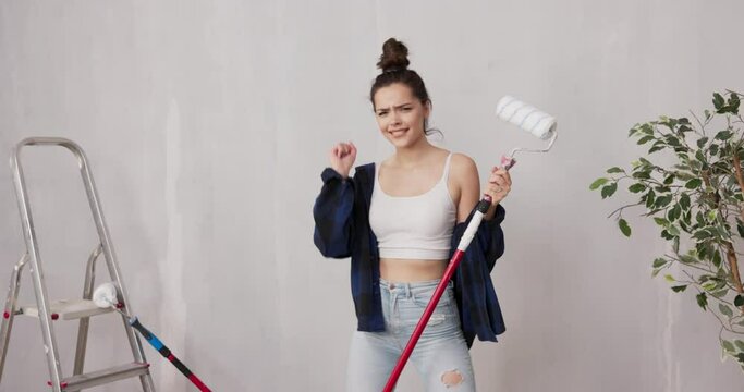 Smiling pretty girl dressed in tight clothing shirt dances with paint roller stick on walls beside her stands ladder, woman spends time in new apartment on finishing touches, renovation