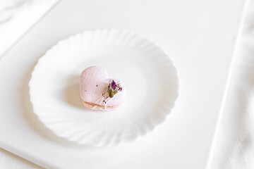 A pink heart-shaped cake lies on a white plate on a light background. Light macaroons on a white canvas. Valentine's Day background