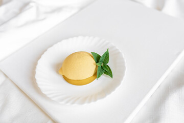 Mousse yellow cake in the form of a lemon with a green mint leaf on a white plate on a light background. Delicious and beautiful pastries. Background for confectionery