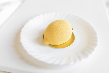 Yellow mousse cake in the form of a lemon lies on a white plate on a light background. Interesting and tasty food. Culinary background